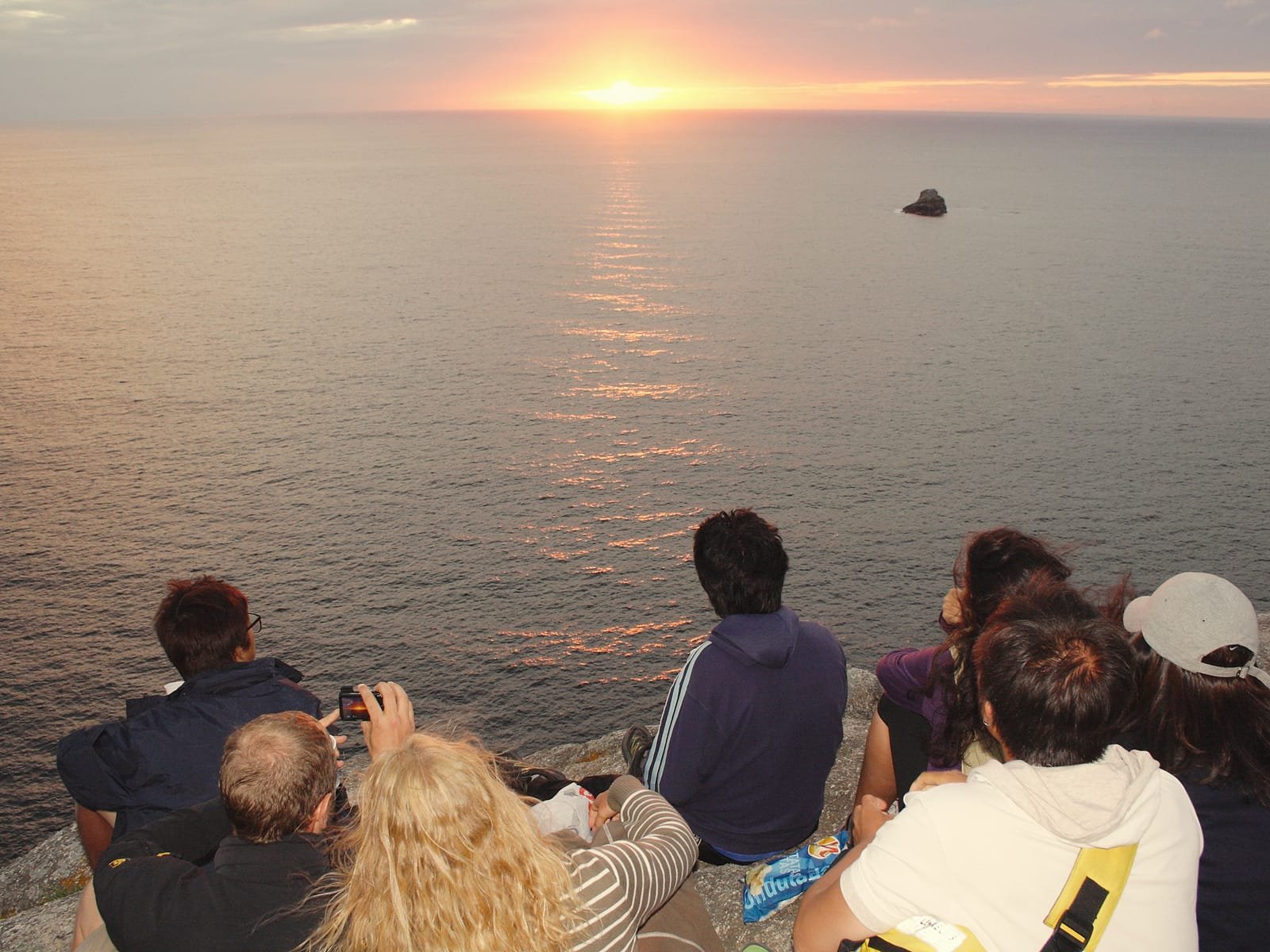 A group of pilgrims sitting together on the rocks of Cape Fisterra, watching the sunset over the Atlantic